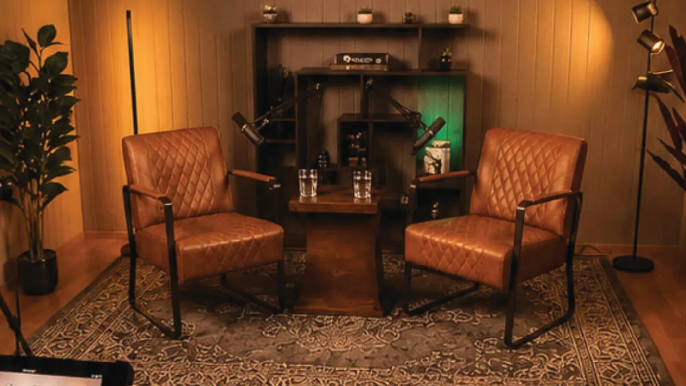 Studio-Set Ember: zwei braune Ledersessel mit Holztisch, Mikrofonen und Bücherregal in warmem Licht