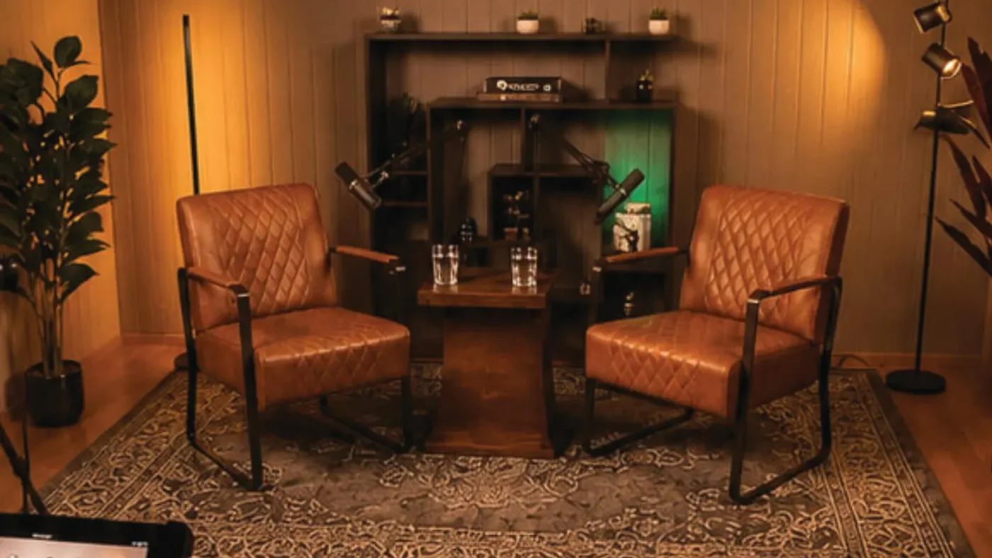 Studio-Set Ember: zwei braune Ledersessel mit Holztisch, Mikrofonen und Bücherregal in warmem Licht